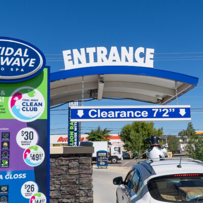 The entrance of Tidal Wave Auto Spa featuring various car wash options on a clear day, with a staff member ready to assist and signage promoting free vacuums with each wash. 